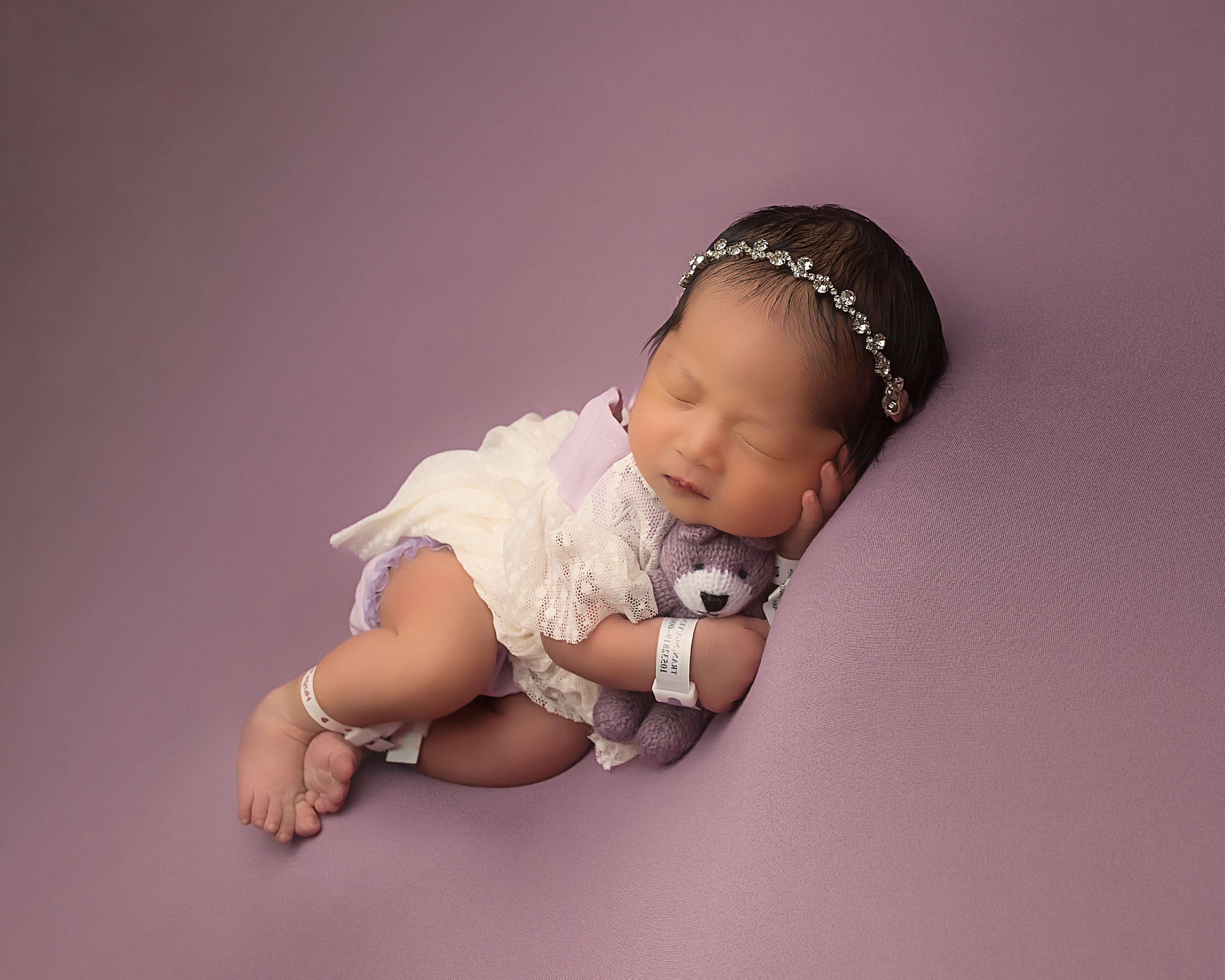 Newborn baby girl sleeping in a lace outfit on a purple backdrop, holding a knit teddy bear